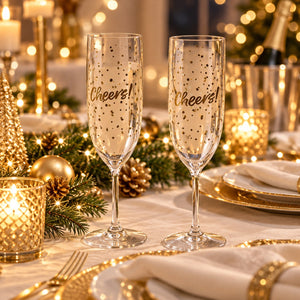 Two clear champagne flutes with gold confetti and the word 'Cheers!' printed on them, set on a decorated holiday table with candles, pine branches, gold ornaments, plates, and warm string lights in the background.