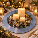 A festive Christmas table centerpiece featuring three cream pillar candles arranged on a round slate blue charger plate, surrounded by pine branches, pinecones, silver and gold ornaments, and a small gold star, with warm string lights in the background.