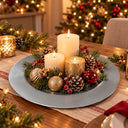 A festive Christmas table centerpiece featuring three lit pillar candles in white and textured gold, surrounded by pinecones, red berries, green pine branches, and gold and red ornaments on a round silver charger plate, set on a decorated dining table with a blurred Christmas tree in the background.