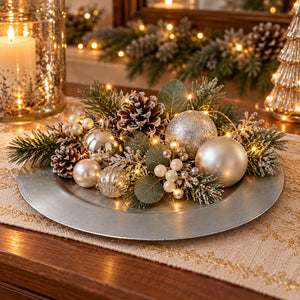A festive holiday centerpiece arranged on a round silver platter, featuring silver and gold ornaments, pinecones, green foliage, frosted greenery, and warm string lights on a wooden table.