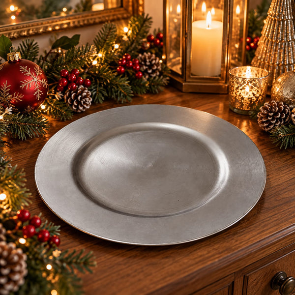 A round silver charger plate with a brushed metal finish, styled on a wooden table with festive Christmas decorations including pine branches, red berries, pinecones, a red ornament, and lit candles.