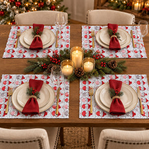 Four matching Christmas-themed placemats arranged on a wooden dining table, each styled with white plates, gold cutlery, a folded red napkin, and holiday greenery and candles in the center.