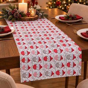 A festive Christmas table runner with a repeating pattern of snowmen in red and gray hats, styled on a wooden dining table with holiday decorations, plates, and candles.