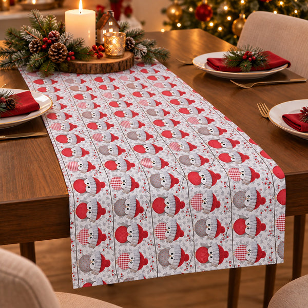A festive Christmas table runner with a repeating pattern of snowmen in red and gray hats, styled on a wooden dining table with holiday decorations, plates, and candles.