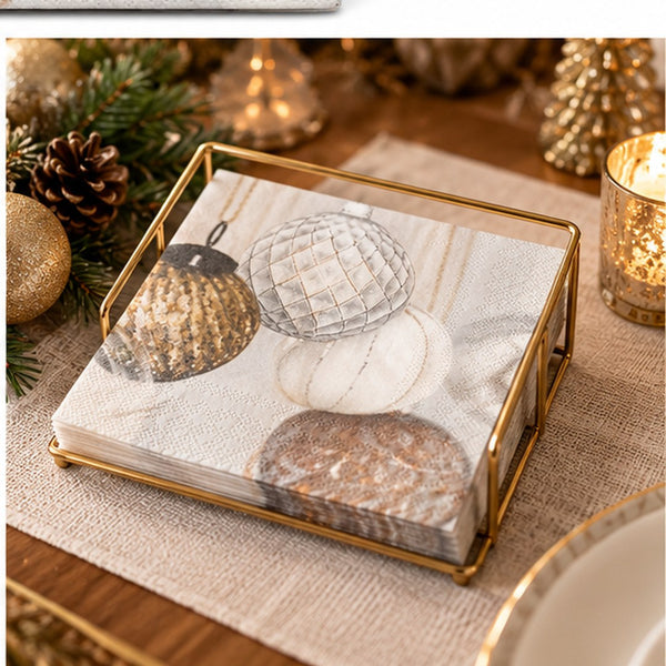 A square gold-framed napkin holder filled with white paper napkins printed with a holiday ornament pattern, styled on a burlap table runner with pine branches, a candle, and gold decorative accents.
