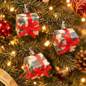 Three Christmas ornament shaped like wrapped gift boxes hang on a decorated Christmas tree. Each box is wrapped in a holly-patterned paper with a red ribbon bow and silver clip hanger.