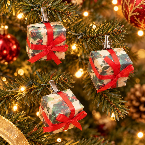 Three Christmas ornament shaped like wrapped gift boxes hang on a decorated Christmas tree. Each box is wrapped in a holly-patterned paper with a red ribbon bow and silver clip hanger.