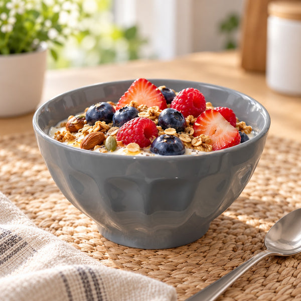 A gray ceramic bowl filled with granola, blueberries, strawberries, and raspberries, set on a woven placemat on a wooden table with a spoon nearby.