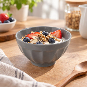 A gray ceramic bowl filled with granola, blueberries, and sliced strawberries on a wooden table, with a spoon and kitchen items softly blurred in the background.