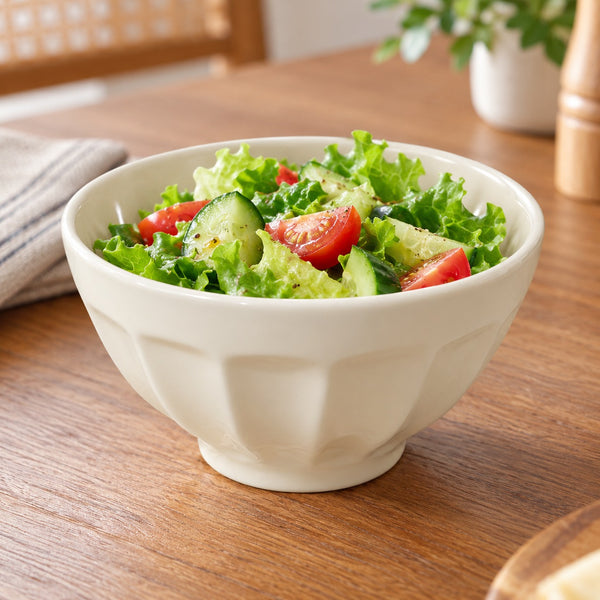 A white ceramic salad bowl filled with green lettuce, cucumber, and red tomato slices, set on a wooden table in a softly blurred kitchen setting.