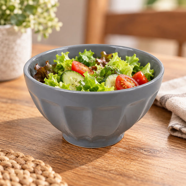 A gray ceramic salad bowl filled with fresh greens, cucumber slices, and cherry tomatoes, set on a wooden table with a blurred background.