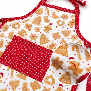 A festive kitchen apron with a white background printed with gingerbread cookies, trees, and candy canes, accented with red polka-dot trim and a large red polka-dot front pocket. A circular inset shows a close-up of the pocket fabric.