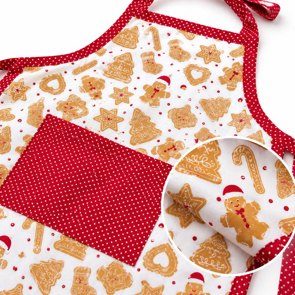 A festive kitchen apron with a white background printed with gingerbread cookies, trees, and candy canes, accented with red polka-dot trim and a large red polka-dot front pocket. A circular inset shows a close-up of the pocket fabric.