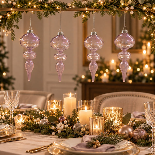 Four lavender glass hanging ornament lights suspended from a decorated Christmas table with candles, greenery, and warm string lights in a cozy indoor setting.
