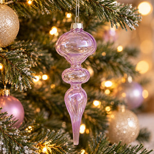 A translucent lavender purple glass Christmas ornament shaped like a teardrop with rounded middle sections, hanging from a decorated Christmas tree with warm lights and other ornaments.
