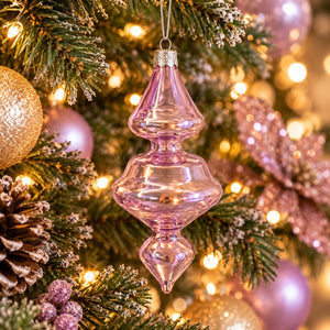 A pink glass Christmas tree ornament hanging from a decorated Christmas tree with warm string lights, pinecones, and gold and pink ornaments in the background.