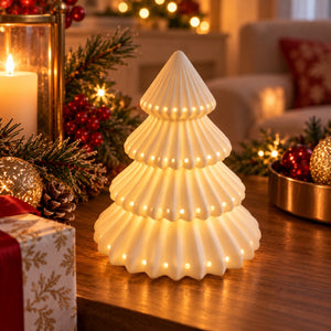 A small illuminated Christmas tree lamp made of pleated white ceramic or resin, glowing warmly on a wooden table in a cozy holiday living room setting with candles, ornaments, and festive decor.