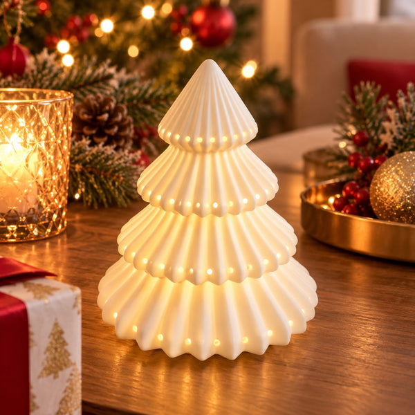 A small white ribbed Christmas tree-shaped table lamp with warm glowing lights, placed on a wooden table in a cozy holiday setting with candles, pine branches, ornaments, and wrapped gift boxes.