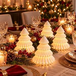 Four white pleated Christmas tree-shaped candle holders with warm glowing lights, arranged on a decorated holiday table with green garlands, red berries, and candles.
