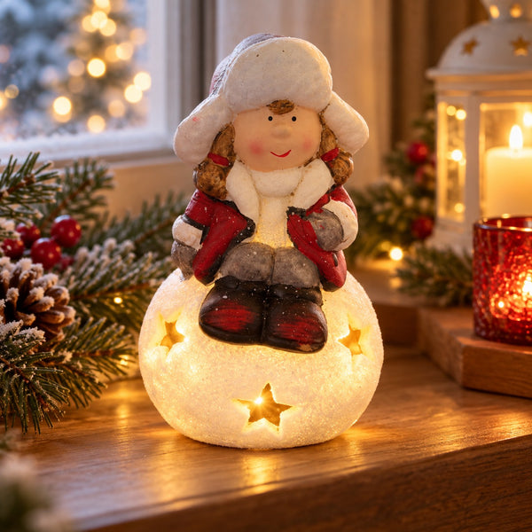 A festive Christmas snowman figurine sitting on a glowing star-lit base, styled on a wooden surface with holiday greenery, a lantern, and warm blurred lights in the background.