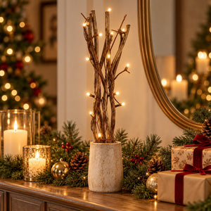 A decorative holiday scene with a central dried twig branch arrangement in a textured cream pot, lit with warm white string lights, placed on a wooden console table with candles, pine garland, ornaments, and wrapped gifts in a warmly lit room.