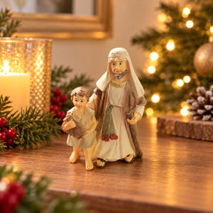 A small decorative figurine of a bearded man in white and brown robes holding a child, displayed on a wooden surface with warm holiday lights, a lit candle, and pine branches in the background.