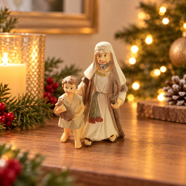 A small decorative figurine of a bearded man in white and brown robes holding a child, displayed on a wooden surface with warm holiday lights, a lit candle, and pine branches in the background.