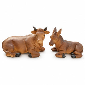Two small brown animal figurines, a cow lying down on the left and a donkey lying down on the right, photographed on a plain white background.