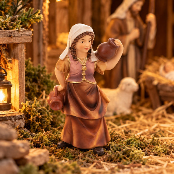 A hand-painted nativity figurine of a woman carrying water jugs, set in a rustic stable scene with warm lighting, a sheep, and a baby crib in the background.