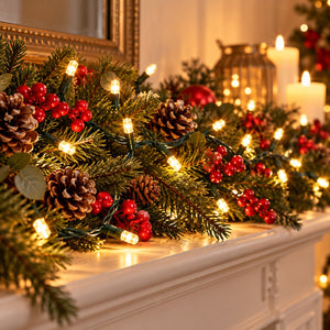 A festive Christmas garland draped across a white fireplace mantel, decorated with green pine branches, brown pinecones, clusters of red berries, and warm white string lights, with candles and a mirror in the softly blurred background.