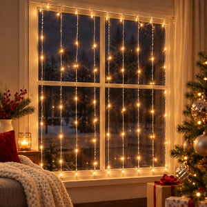 Warm white curtain string lights draped across a four-pane window, creating a cozy holiday atmosphere in a living room setting with a Christmas tree and wrapped gifts visible.