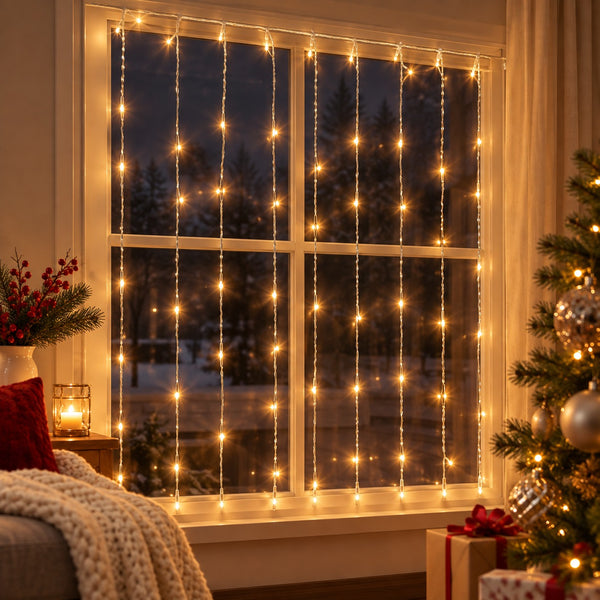 Warm white curtain string lights draped across a four-pane window, creating a cozy holiday atmosphere in a living room setting with a Christmas tree and wrapped gifts visible.