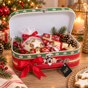 A red and green vintage-style metal gift box with a hinged lid, decorated with a red ribbon bow and a hanging 'noah' tag, filled with holiday treats including star-shaped cookies, wrapped gifts, ornaments, pinecones, and evergreen branches on a wooden table.
