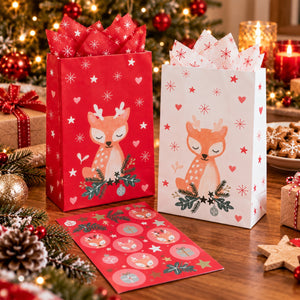 Two Christmas-themed paper gift bags and matching sticker cards on a wooden table, surrounded by holiday decorations including lights, ornaments, pine branches, and cookies. One bag is red with a white deer illustration and snowflake pattern, and the other is white with a red deer illustration and heart pattern.