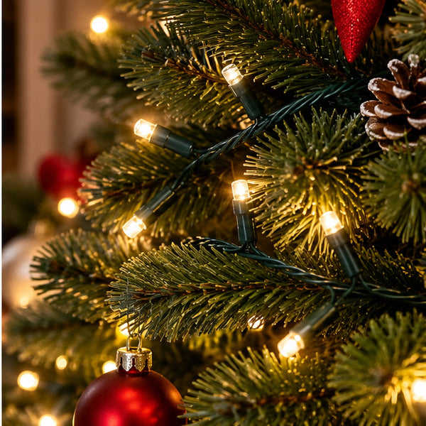 Warm white Christmas tree string lights wrapped around green pine branches, with a red ornament and pinecone visible in a cozy holiday setting.