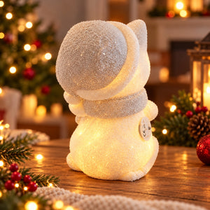 A glittery snowman figurine glowing with warm white light, wearing a matching glittery hat and scarf, set on a wooden table in a cozy Christmas-themed room with blurred lights and decorations in the background.