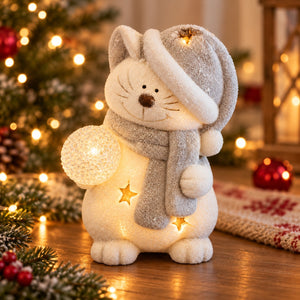 A festive white cat Christmas ornament wearing a glittery gray hat and scarf, holding a glowing orb, with warm star lights inside, set on a wooden surface in a decorated holiday room.