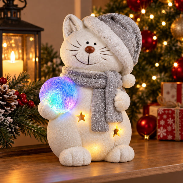 A white fluffy cat Christmas ornament or figurine wearing a gray hat and scarf, holding a glowing multicolor orb, with warm star lights on its body, displayed on a wooden surface in a festive holiday setting.
