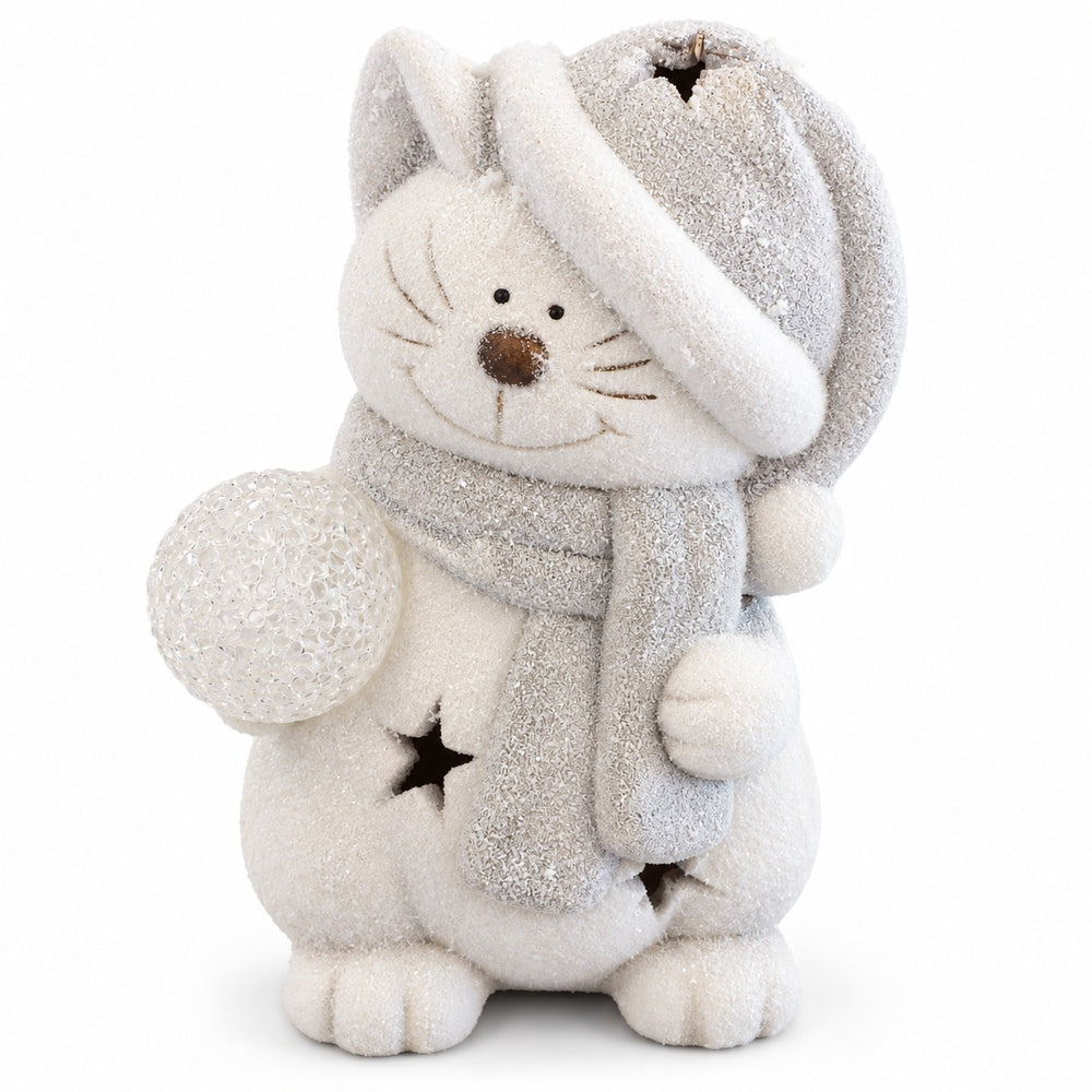 A white fluffy cat Christmas ornament or figurine wearing a gray hat and scarf, holding a glowing multicolor orb, with warm star lights on its body, displayed on a wooden surface in a festive holiday setting.