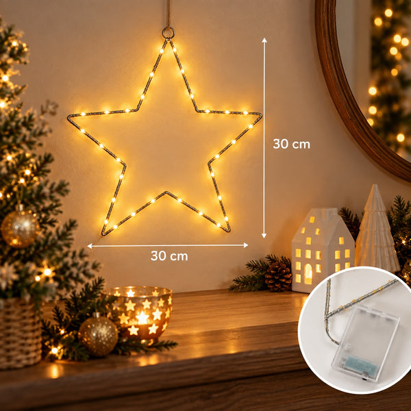 A glowing star-shaped Christmas string light hangs on a wall above a wooden mantel, with dimension arrows showing 30 cm width and 30 cm height. The scene includes a decorated tree, small house and cone ornaments, pinecones, and a circular inset showing the light in its clear hanging case.