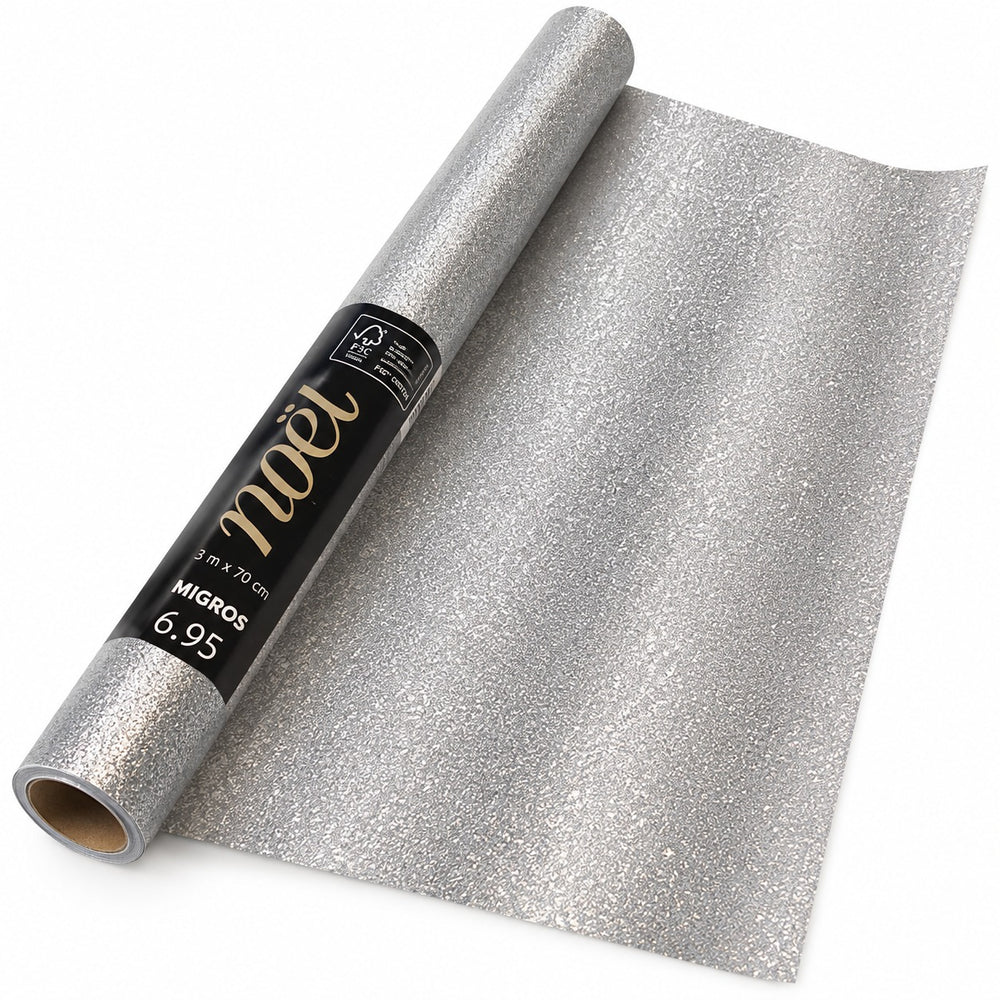 A roll of sparkly silver glitter wrapping paper is unrolled beside a wrapped gift box with a matching ribbon and bow, styled with Christmas greenery, pinecones, red berries, and warm holiday lights on a wooden surface.