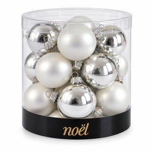 A clear cylindrical container filled with assorted silver and white Christmas ornaments, with a black band at the bottom printed with the gold 'noél' brand name.
