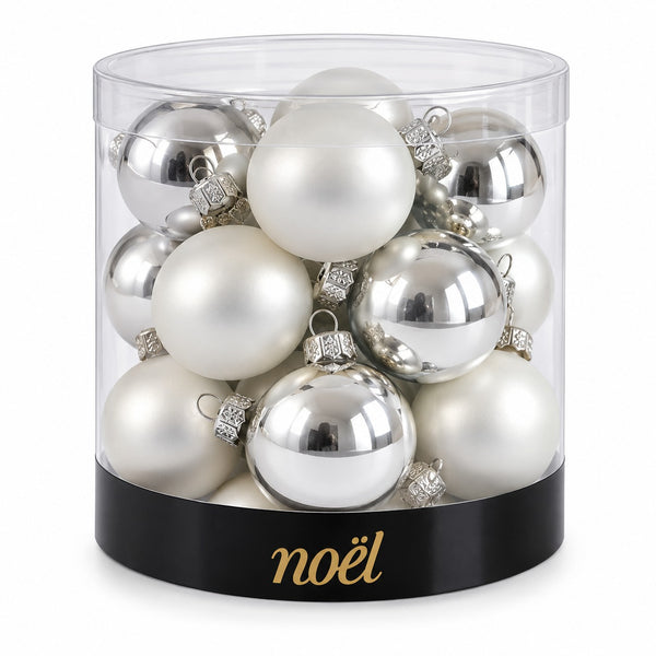 A clear cylindrical container filled with assorted silver and white Christmas ornaments, with a black band at the bottom printed with the gold 'noél' brand name.