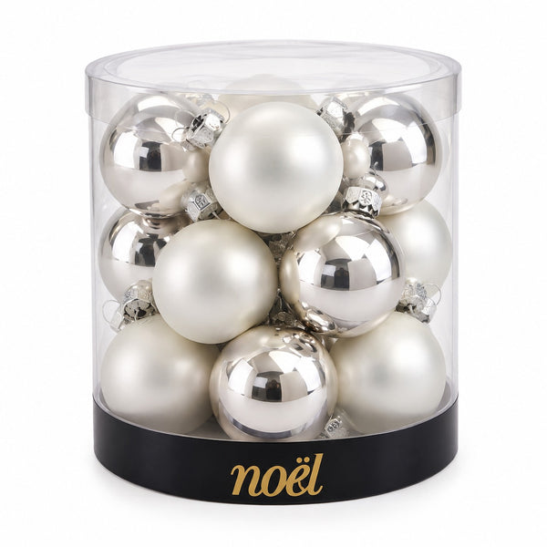 A set of 12 Christmas ornaments in a clear cylindrical container with a black base printed with the brand name 'noël' in gold. The ornaments are a mix of shiny silver and matte off-white/ivory spheres.