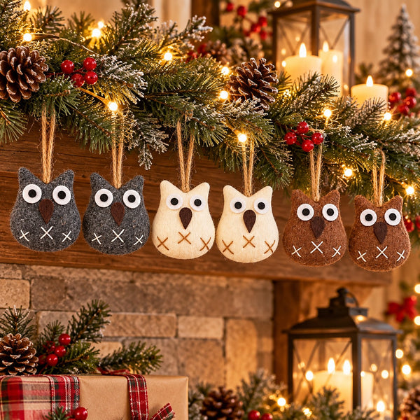 Six felt owl Christmas ornaments in gray, cream, and brown hang from a decorated wooden mantel with pine branches, candles, and warm string lights.