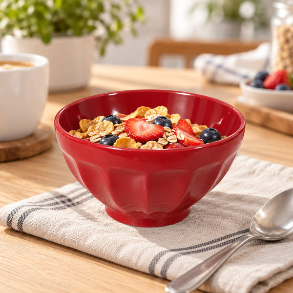 A red ceramic bowl filled with cereal, blueberries, and sliced strawberries, set on a beige striped cloth on a wooden table with a spoon nearby.