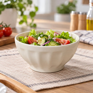 A white ceramic salad bowl filled with fresh greens, tomatoes, cucumbers, and red onion, set on a woven placemat on a wooden table with blurred kitchen items in the background.