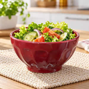 A glossy dark red ceramic salad bowl filled with lettuce, cucumber, tomato, and red onion, placed on a woven beige placemat in a softly blurred kitchen setting.