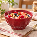 A glossy red ceramic bowl filled with yogurt, granola, strawberries, and blueberries, set on a woven placemat with a spoon on a wooden table in a softly blurred kitchen setting.