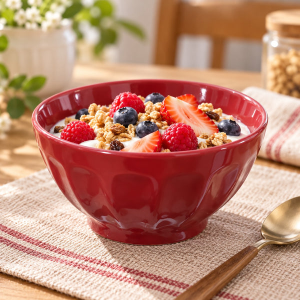 A glossy red ceramic bowl filled with yogurt, granola, strawberries, and blueberries, set on a woven placemat with a spoon on a wooden table in a softly blurred kitchen setting.